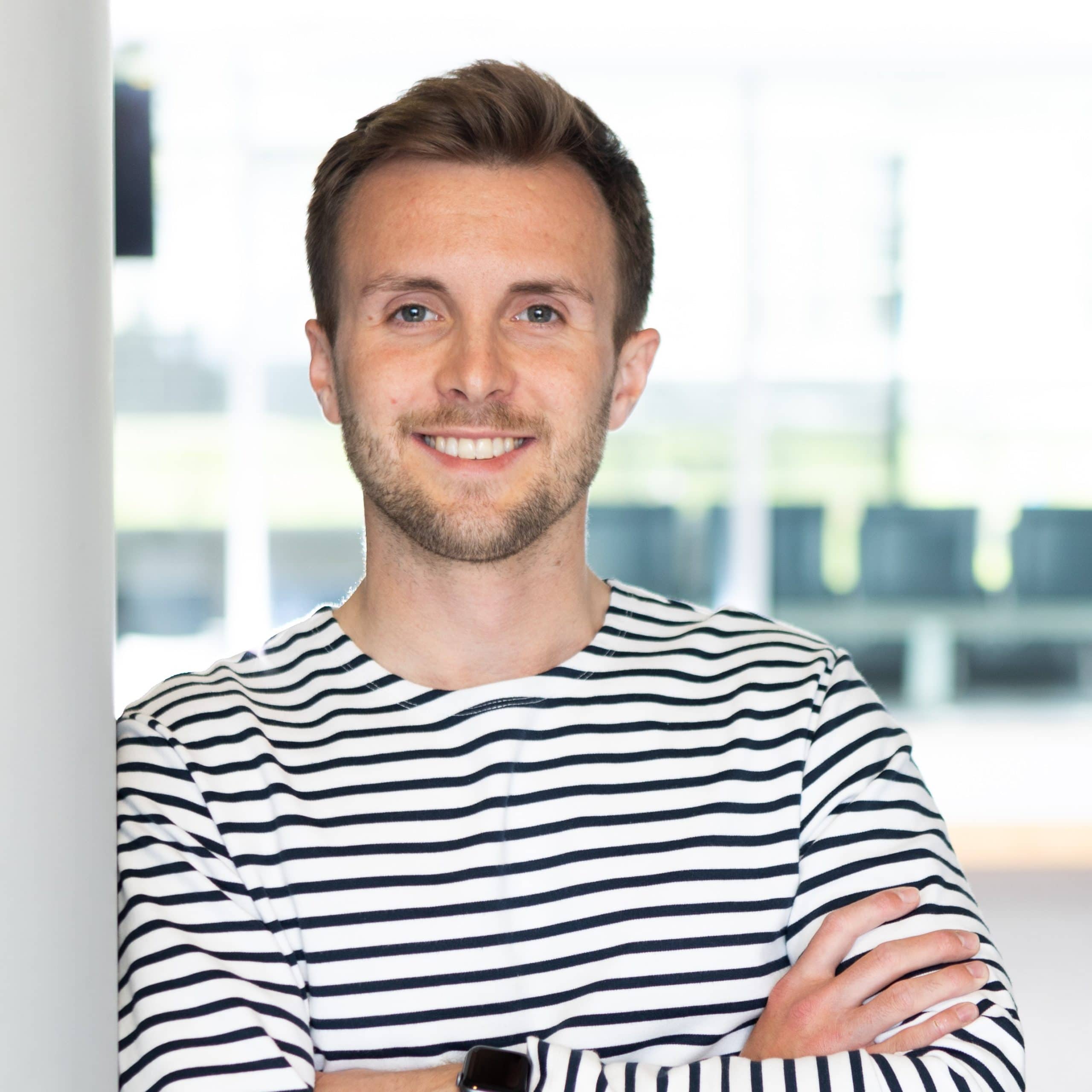 Matthew Regan a white masculine person with light brown hair and beard crossing their arms and smiling in a stripey black and white top.