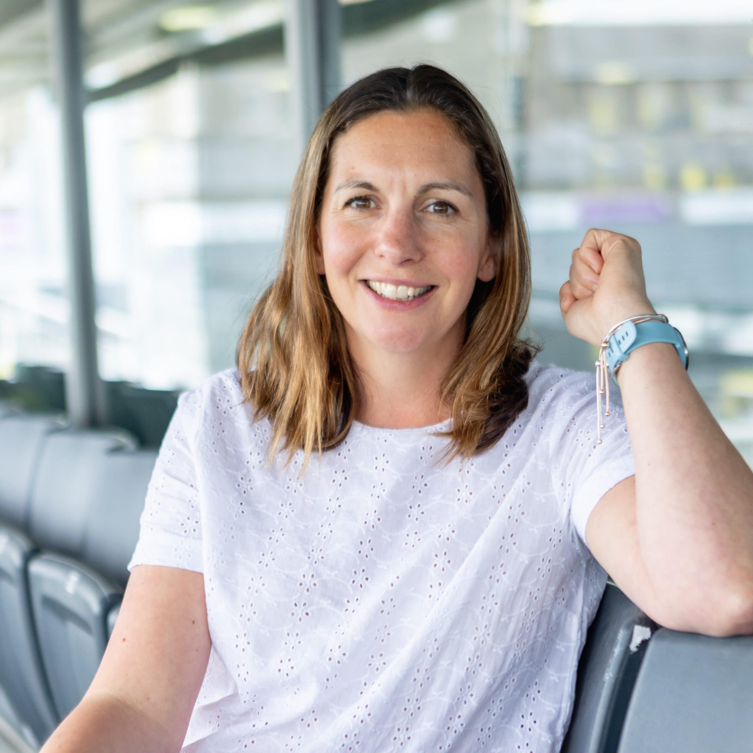 A white feminine person with shoulder length blonde-brown hair wearing a white top and blue watch.