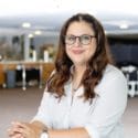 A brunette feminine person with curly long hair, wearing a white shirt and black glasses.