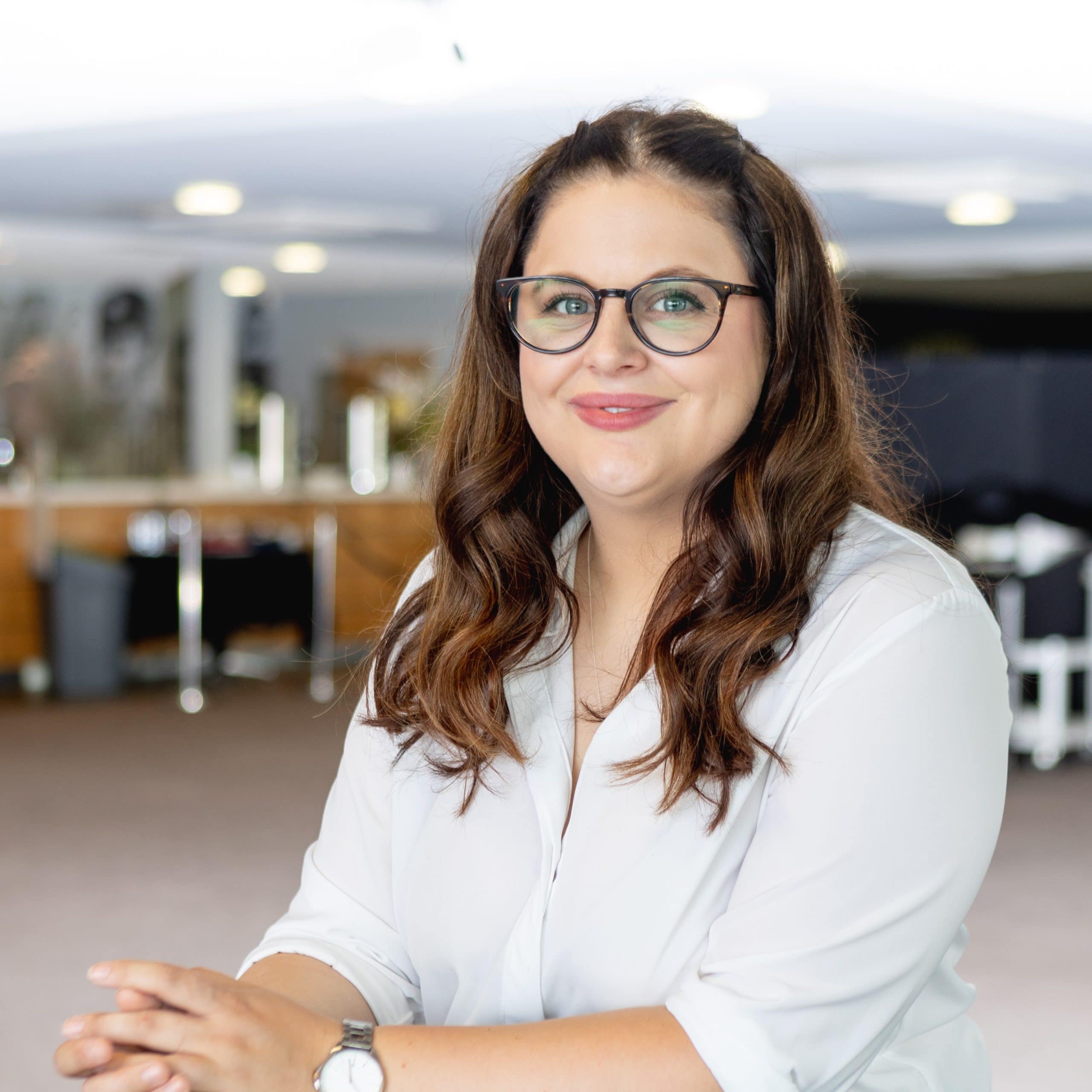 Lauren Roberts A brunette feminine person with curly long hair, wearing a white shirt and black glasses.