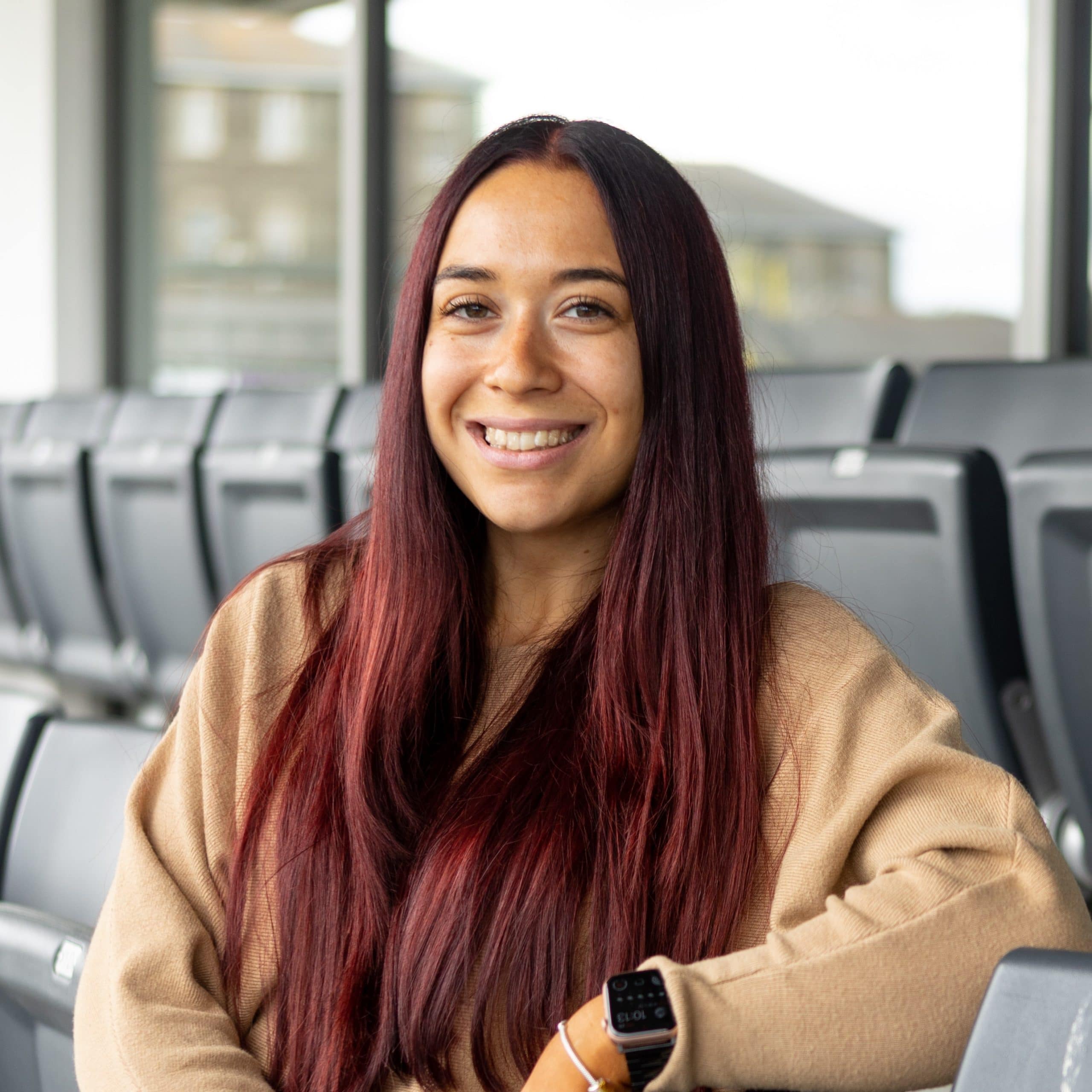 A feminine person with very long maroon coloured hair, tanned skin, wearing a light brown jumper and an apple watch.