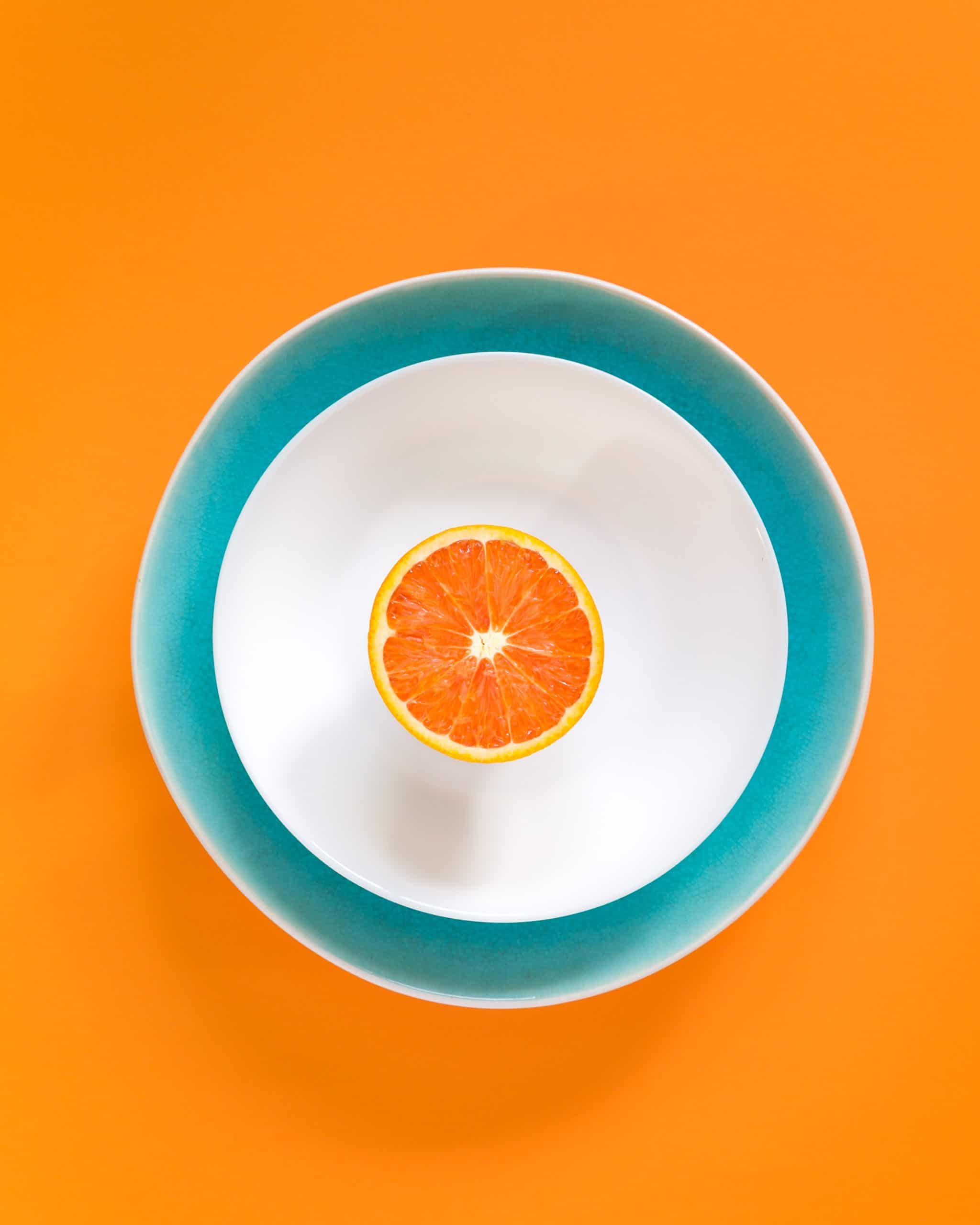 half of an orange in a white and teal bowl on an orange background.